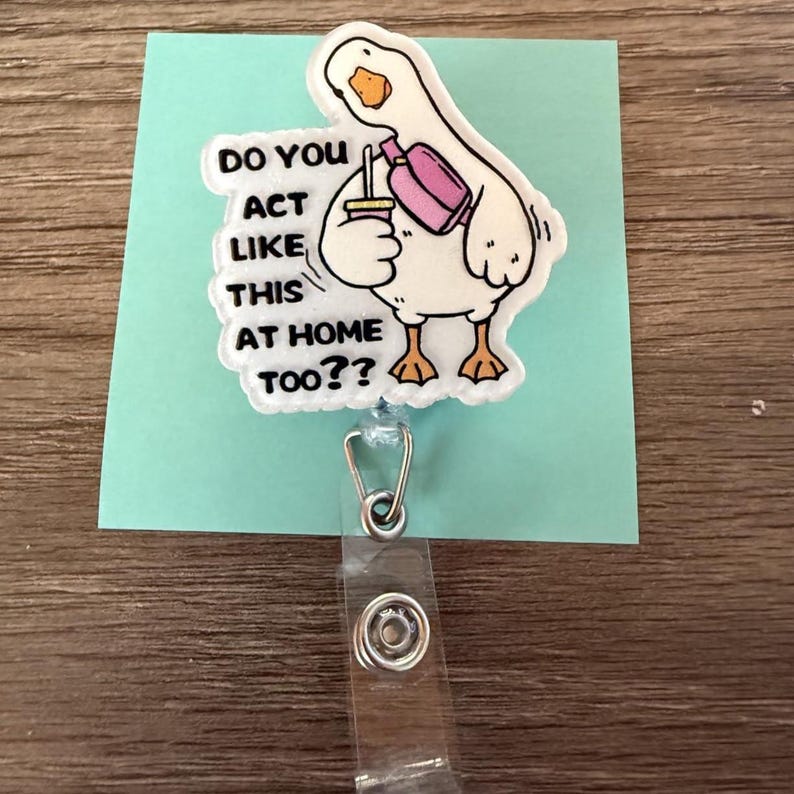 K&ouml;nnte beinhalten: Ein wei&szlig;es G&auml;nse-Badge-Rolle mit dem Text "DO YOU ACT LIKE THIS AT HOME TOO??" Die Gans h&auml;lt ein Getr&auml;nk und eine Flasche. Die Badge-Rolle ist an einer hellblauen Karte befestigt.