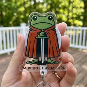 May include: A clear acrylic keyring featuring a cartoon frog in a red cape, holding a sword. The frog has a serious expression. The text "I'M BURNT OUT M'LORD" is printed at the bottom. A silver keyring is attached.