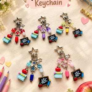 May include: Five metal star-shaped keychains featuring charms of miniature books, red apples, pencils, and black chalkboards with the text ABC. Each keychain includes colorful beads and star accents, arranged in a display with the text Teacher Keychain.
