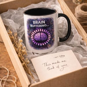 May include: White ceramic mug with a black handle and rim, featuring the text "BRAIN BUFFERING..." above a purple brain graphic. The mug is in a gift box with a handwritten note that says "This made me think of you."