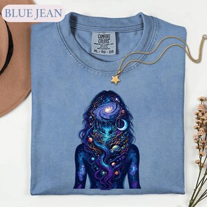 May include: A blue jean colored t-shirt with a Comfort Colors label in the neck. The shirt features a graphic of a silhouette filled with a cosmic scene of a galaxy, planets, a crescent moon, and a forest landscape with flowing blue hair.