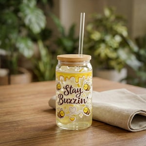 Puede incluir: Un vaso de vidrio transparente con tapa de madera y pajita de vidrio. El vaso presenta un diseño amarillo con abejas y el texto "Stay Buzzin'". El vaso está lleno de líquido amarillo y hielo.
