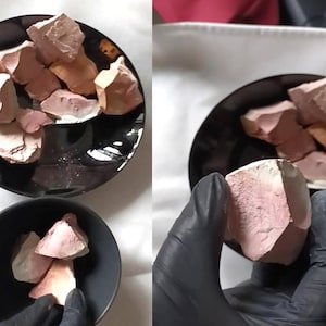 May include: A collection of raw, earthy-toned clay chunks in a black bowl. The clay pieces vary in size and shape, with a mix of pink, beige, and white hues. Some pieces are held by a gloved hand. The image suggests a natural product.