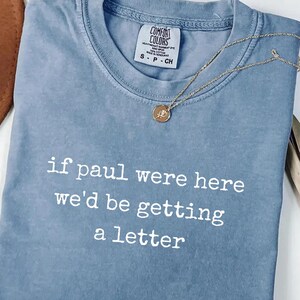 May include: Blue jean Comfort Colors t-shirt with the text "if paul were here we'd be getting a letter" in white. A gold necklace with a pendant is draped across the shirt. The shirt is folded on a white surface.