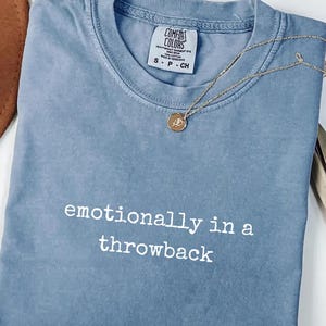 Könnte beinhalten: Ein blaues Comfort Colors T-Shirt mit dem Text "emotional in einem Throwback" in Weiß. Eine goldene Halskette mit Anhänger liegt über dem Shirt. Das Shirt ist auf einer weißen Oberfläche gefaltet.