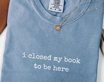 Könnte beinhalten: Ein blaues T-Shirt in Jeansoptik mit Rundhalsausschnitt und dem weißen Schriftzug "i closed my book to be here" auf der Vorderseite. Das Shirt hat ein Comfort Colors Markenetikett am Innenkragen und wird mit einer goldfarbenen Halskette kombiniert.