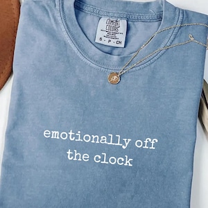 May include: A blue jean Comfort Colors t-shirt with the text "emotionally off the clock" in white. A gold necklace with a pendant is draped across the shirt. The shirt is folded on a white surface.