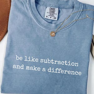 May include: A blue jean coloured short-sleeve t-shirt with a crew neckline. The shirt features the white text "be like subtraction and make a difference" printed on the front. A gold-toned necklace with a small circular pendant rests on the fabric.