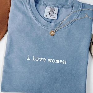May include: A blue jean Comfort Colors t-shirt with the text "i love women" in white. A gold necklace with a pendant is draped across the shirt. The shirt is folded and displayed on a white surface.