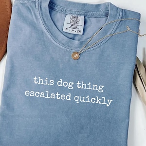 May include: A blue jean colored t-shirt with a crew neckline and the white text "this dog thing escalated quickly" printed on the front. The shirt features a Comfort Colors brand label at the inner neck and is paired with a gold tone pendant necklace.
