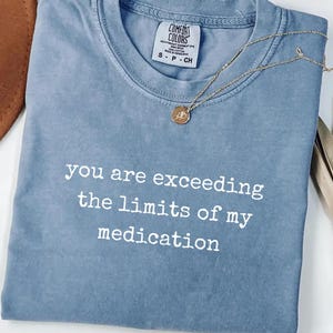 Może przedstawiać: Niebieska koszulka z okrągłym dekoltem i białym napisem "you are exceeding the limits of my medication" nadrukowanym z przodu. Koszulka posiada metkę marki Comfort Colors przy wewnętrznej stronie kołnierza oraz krótki rękaw.