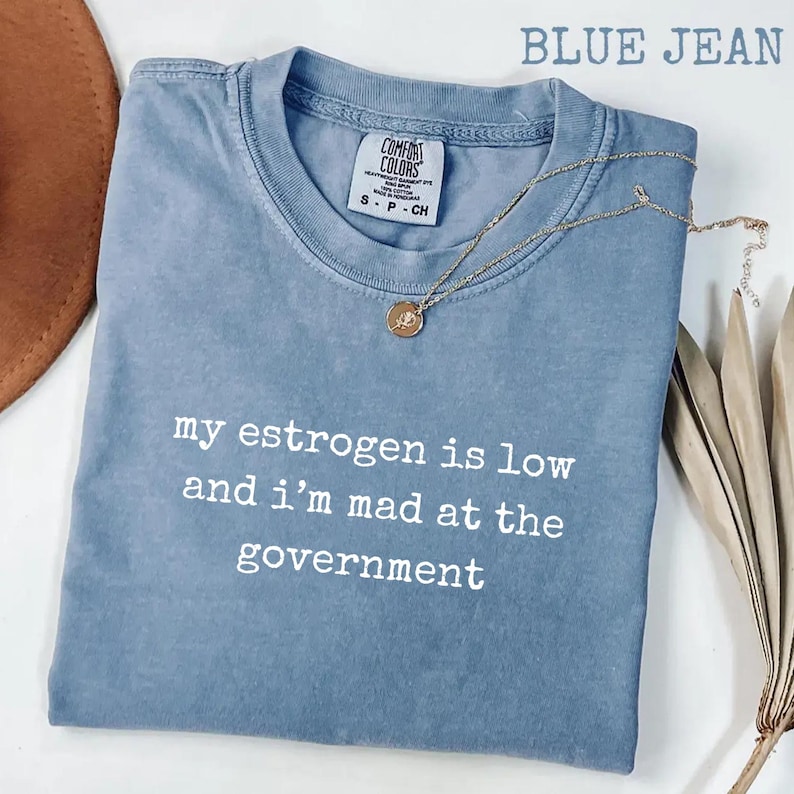 K&ouml;nnte beinhalten: Ein kurz&auml;rmeliges T-Shirt in Jeansblau mit Rundhalsausschnitt. Das Shirt ist mit wei&szlig;em Text bedruckt, der lautet, my estrogen is low and i'm mad at the government. Eine goldfarbene Halskette mit einem kleinen runden Anh&auml;nger liegt auf dem Stoff.