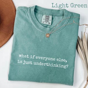 May include: A light green short-sleeve t-shirt with a crew neckline and a Comfort Colors brand label at the inner collar. The shirt features the white printed text, what if everyone else, is just underthinking? A thin silver chain necklace is present.
