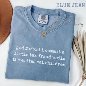 Op de afbeelding: Een spijkerblauw t-shirt met korte mouwen en een ronde hals. Het shirt heeft witte tekst op de voorkant gedrukt die luidt: god forbid i commit a little tax fraud while the elites eat children. Een goudkleurige ketting rust op de kraag van het shirt.