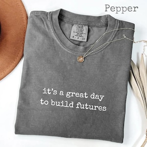 Puede incluir: Una camiseta gris Comfort Colors con el texto "it's a great day to build futures" en blanco. Un collar plateado con un peque&ntilde;o colgante est&aacute; sobre la camiseta. La palabra "Pepper" est&aacute; en la parte superior de la imagen.