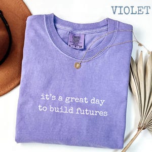 Puede incluir: Camiseta violeta Comfort Colors con el texto blanco "it's a great day to build futures". Un collar dorado con un colgante descansa sobre la camiseta. La camiseta est&aacute; doblada y se muestra con un sombrero marr&oacute;n y hojas de palma secas.