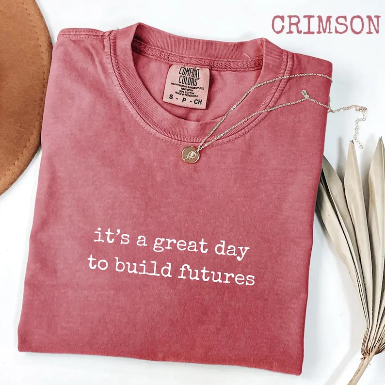 Puede incluir: Camiseta de color carmes&iacute; con el texto blanco "it's a great day to build futures". Un collar plateado con un colgante descansa sobre la camiseta. La camiseta es de Comfort Colors.