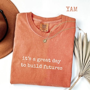 Puede incluir: Una camiseta Comfort Colors de color coral con la frase blanca "it's a great day to build futures". Un collar dorado con un colgante descansa sobre la camiseta. Un sombrero marr&oacute;n y una planta seca est&aacute;n en el fondo.