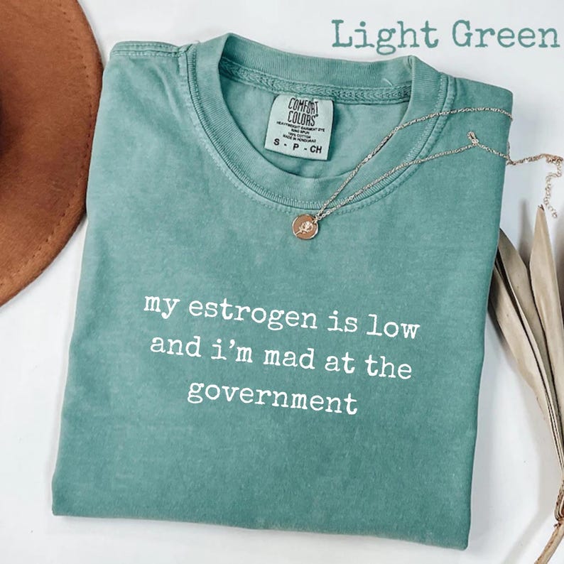 K&ouml;nnte beinhalten: Ein hellgr&uuml;nes Kurzarm-T-Shirt mit dem wei&szlig;en aufgedruckten Text "my estrogen is low and i'm mad at the government" auf der Vorderseite. Das Kleidungsst&uuml;ck hat einen Rundhalsausschnitt und ein Comfort Colors Markenetikett im inneren Kragenbereich.
