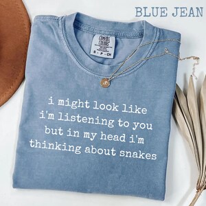 Könnte beinhalten: Ein kurzärmeliges T-Shirt in Jeansblau mit Rundhalsausschnitt und einem weißen grafischen Aufdruck, der lautet: I might look like i'm listening to you but in my head i'm thinking about snakes. Eine goldfarbene Halskette mit einem kleinen runden Anhänger liegt darauf.