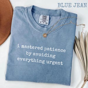 May include: A blue jean colored t-shirt with a crew neckline and a Comfort Colors label. The shirt features white text that reads, I mastered patience by avoiding everything urgent. A gold-toned necklace with a small circular pendant rests on top.