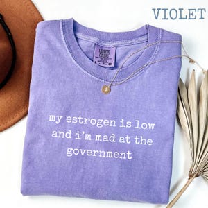 K&ouml;nnte beinhalten: Ein violettes kurz&auml;rmeliges T-Shirt mit Rundhalsausschnitt und dem wei&szlig;en Aufdruck 'my estrogen is low and i'm mad at the government'. Das T-Shirt hat ein Comfort Colors-Etikett am Innenkragen und wird zusammen mit einer goldfarbenen Kette gezeigt.