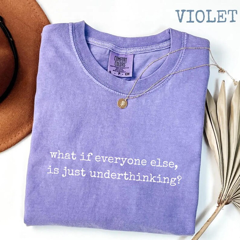 May include: A violet short-sleeved t-shirt with a crew neckline and a Comfort Colors label. The shirt features the white printed text, what if everyone else, is just underthinking? A gold-toned chain necklace with a small circular pendant rests on top.