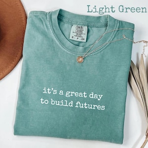 Puede incluir: Camiseta verde claro con el texto blanco "it's a great day to build futures". La camiseta tiene cuello redondo y mangas cortas. Un collar plateado con un colgante est&aacute; sobre la camiseta.