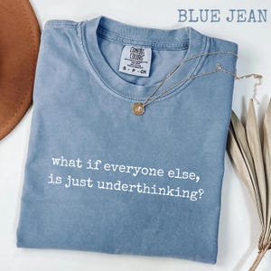 May include: A blue jean colored short-sleeve t-shirt with a crew neckline. The shirt features the white printed text "what if everyone else, is just underthinking?" across the chest. A gold-toned necklace with a small circular pendant rests on top.