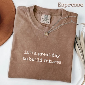 Puede incluir: Camiseta color espresso con el texto "it's a great day to build futures" en blanco. La camiseta tiene cuello redondo y mangas cortas. Un collar plateado con un peque&ntilde;o colgante descansa sobre la camiseta. La camiseta es de Comfort Colors.