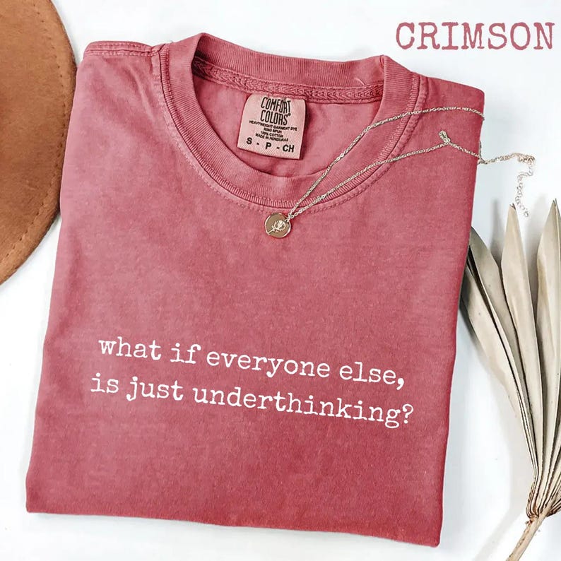 May include: A crimson short-sleeve t-shirt with a crew neckline and the white text "what if everyone else, is just underthinking?" printed on the front. The shirt features a Comfort Colors brand label inside the collar and a silver necklace.
