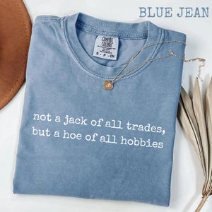 Op de afbeelding: Een spijkerblauw t-shirt met korte mouwen en een ronde hals. Het shirt is voorzien van de witte opdruk, not a jack of all trades, but a hoe of all hobbies. Een goudkleurige ketting met een kleine ronde hanger rust op de kraag.