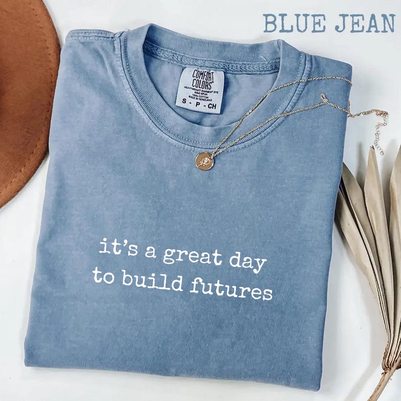 Puede incluir: Una camiseta azul vaquero con el texto "it's a great day to build futures" en blanco. Un collar dorado con un colgante circular descansa sobre la camiseta. La camiseta est&aacute; hecha de un material suave.