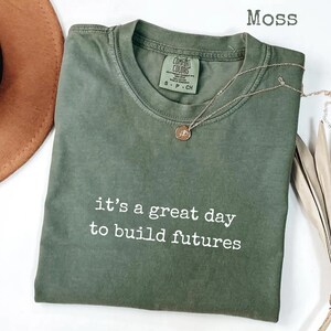 Puede incluir: Una camiseta verde musgo con el texto blanco "it's a great day to build futures". Un collar dorado con un colgante est&aacute; drapeado sobre la camiseta. La camiseta est&aacute; doblada y tiene una etiqueta que dice "Comfort Colors".