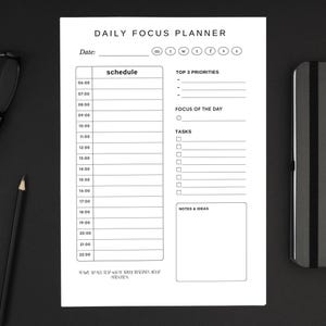 Può includere: Un Daily Focus Planner bianco con sezioni per la pianificazione, le priorità e i compiti. Il planner include il testo "DAILY FOCUS PLANNER". Una matita nera, occhiali e un taccuino sono su una superficie nera.