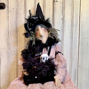 Puede incluir: Una muñeca de tela disfrazada de bruja, con un sombrero negro puntiagudo con plumas, un vestido rosa pálido con dobladillo de encaje y leggings con estampado de leopardo. La muñeca sostiene un bolso negro adornado con flores oscuras y zapatos verde lima.