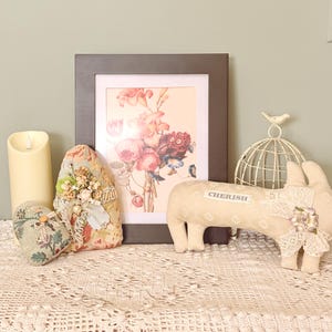 May include: A decorative display featuring a framed floral print, a cream-colored candle, and two fabric heart-shaped pillows. A beige pig-shaped plush toy with the word "Cherish" is also present, along with a decorative birdcage.