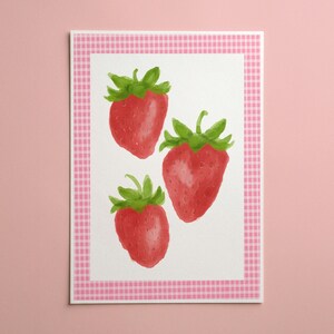 Può includere: Una stampa rettangolare che raffigura tre fragole rosse con foglie verdi su sfondo bianco, incorniciata da un bordo a quadretti vichy rosa e bianco. L'illustrazione mostra i frutti in una disposizione verticale con semi visibili sulla buccia.