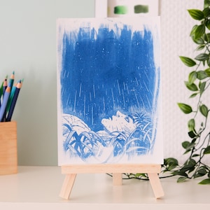 Peut inclure: Une impression d'art rectangulaire présentant une illustration à l'encre bleue d'une personne allongée dans de hautes herbes, regardant un ciel pluvieux. L'œuvre est exposée sur un petit chevalet en bois, révélant des traits texturés.