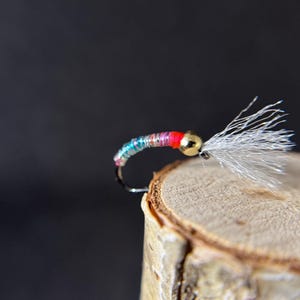May include: A colorful fishing fly with a gold bead head and white feather tail. The fly's body is wrapped with iridescent thread in shades of blue, green, red, and pink. The fly is positioned on a wooden surface against a dark background.