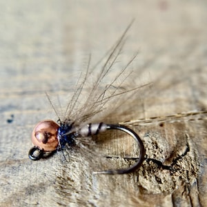May include: Close-up of a fly fishing lure. The lure features a copper-colored bead head, a black hook, and gray feathers. The fly is designed for fishing. The background is a weathered wooden surface.