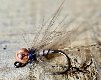 May include: Close-up of a fly fishing lure. The lure features a copper-colored bead head, a black hook, and gray feathers. The fly is designed for fishing. The background is a weathered wooden surface.