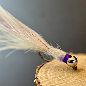 May include: Close-up of a handmade fishing fly. The fly features a silver bead head, a white body with pink and purple accents, and a purple thread wrap. The fly is mounted on a silver hook.