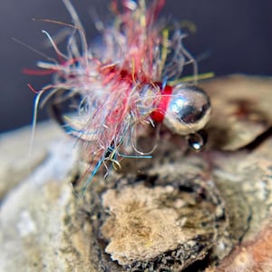 May include: Close-up of a colorful fishing fly. The fly features a silver bead head, red thread, and a vibrant mix of red, blue, yellow, and white fibers. The fly is set against a textured, light brown surface.