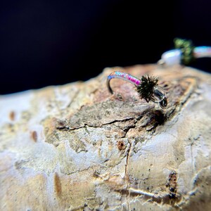 May include: A fishing fly lure featuring a curved metal hook, a translucent body with a vibrant pink and blue iridescent core, and a small dark green fuzzy hackle near the eye of the hook. The lure is positioned on a textured, light-colored surface.