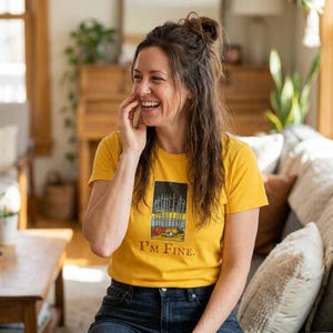 Puede incluir: Camiseta de color amarillo mostaza con un diseño de carta del tarot y el texto "I'M FINE". El diseño de la carta del tarot incluye espadas. La camiseta se combina con vaqueros azules.