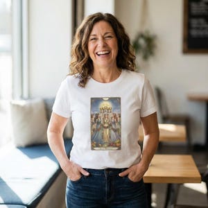 May include: White t-shirt featuring a detailed illustration with figures, a central female figure, and the text "Sacred Wisdom". The artwork has a vintage style with a warm color palette. The shirt is worn by a person.