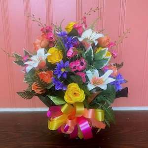 Pode incluir: Um arranjo floral vibrante com uma variedade de flores coloridas, incluindo lírios brancos, rosas amarelas e flores rosa e roxas. O buquê é acentuado com folhagem verde e uma fita multicolorida. O arranjo está sobre um fundo rosa.