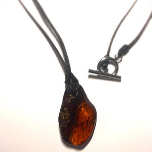 Op de afbeelding: Een hangerketting met een gepolijste barnsteen, met een donkerbruine en oranje kleur. De barnsteen hangt aan een zwart koord met een tuimelsluiting. De ketting wordt gepresenteerd tegen een witte achtergrond.