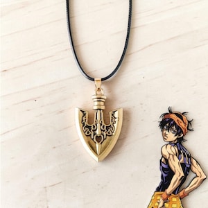 May include: Gold-colored pendant necklace featuring an arrow-shaped design with intricate detailing. The pendant hangs from a black cord. A cartoon character is visible in the background. The necklace is likely a piece of jewelry.